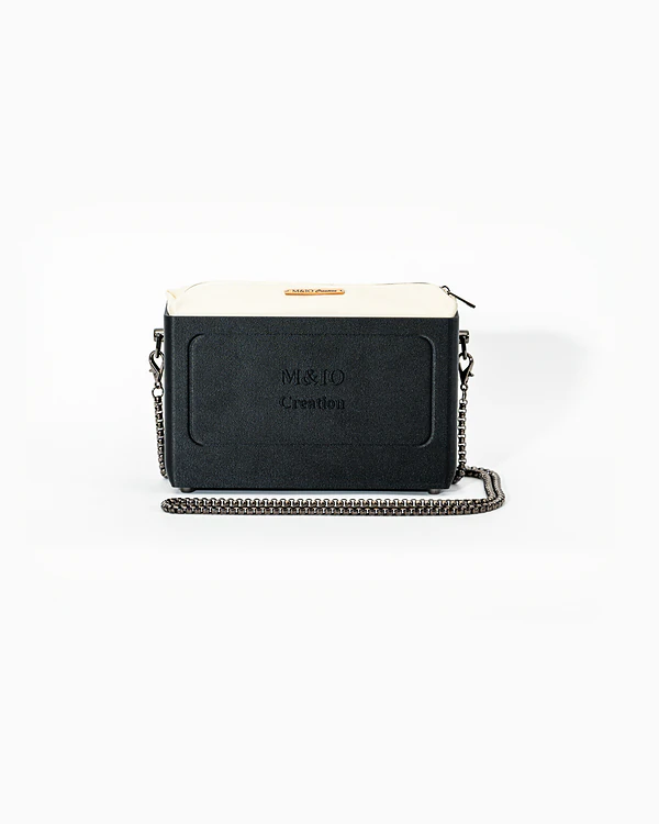 Borsa NYRA di tre quarti: scocca black stone opaca con la scritta M&IO Creation in rilievo sul fronte, pochette bianco panna che sporge dal bordo superiore, catena in metallo canna di fucile.