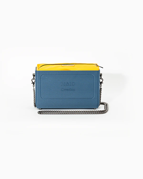 Borsa NYRA di tre quarti: scocca blu portofino opaca con la scritta M&IO Creation in rilievo sul fronte, pochette giallo che sporge dal bordo superiore, catena in metallo canna di fucile.