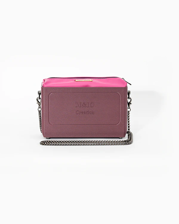 Borsa NYRA di tre quarti: scocca rosso tropea opaca con la scritta M&IO Creation in rilievo sul fronte, pochette fucsia che sporge dal bordo superiore, catena in metallo canna di fucile.