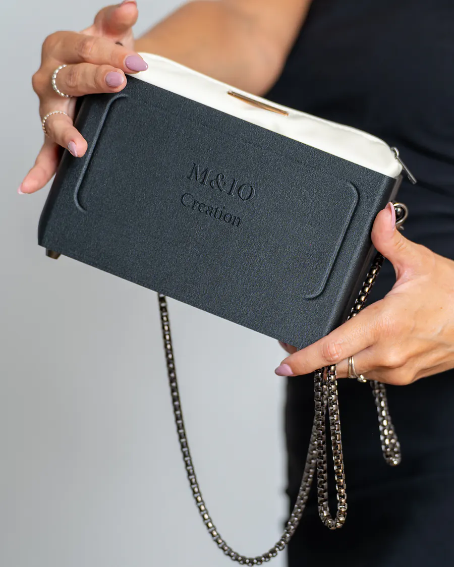 Due mani tengono la borsa Black Stone inclinata, con la scritta M&IO Creation in rilievo; dal bordo esce la pochette bianco panna, la catena pende sotto.