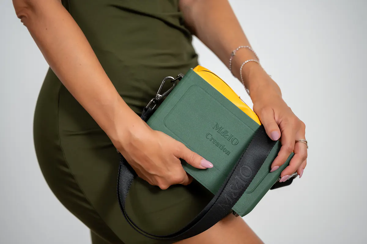 La borsa verde salvia tenuta a due mani all’altezza della vita, con la pochette gialla che sporge.