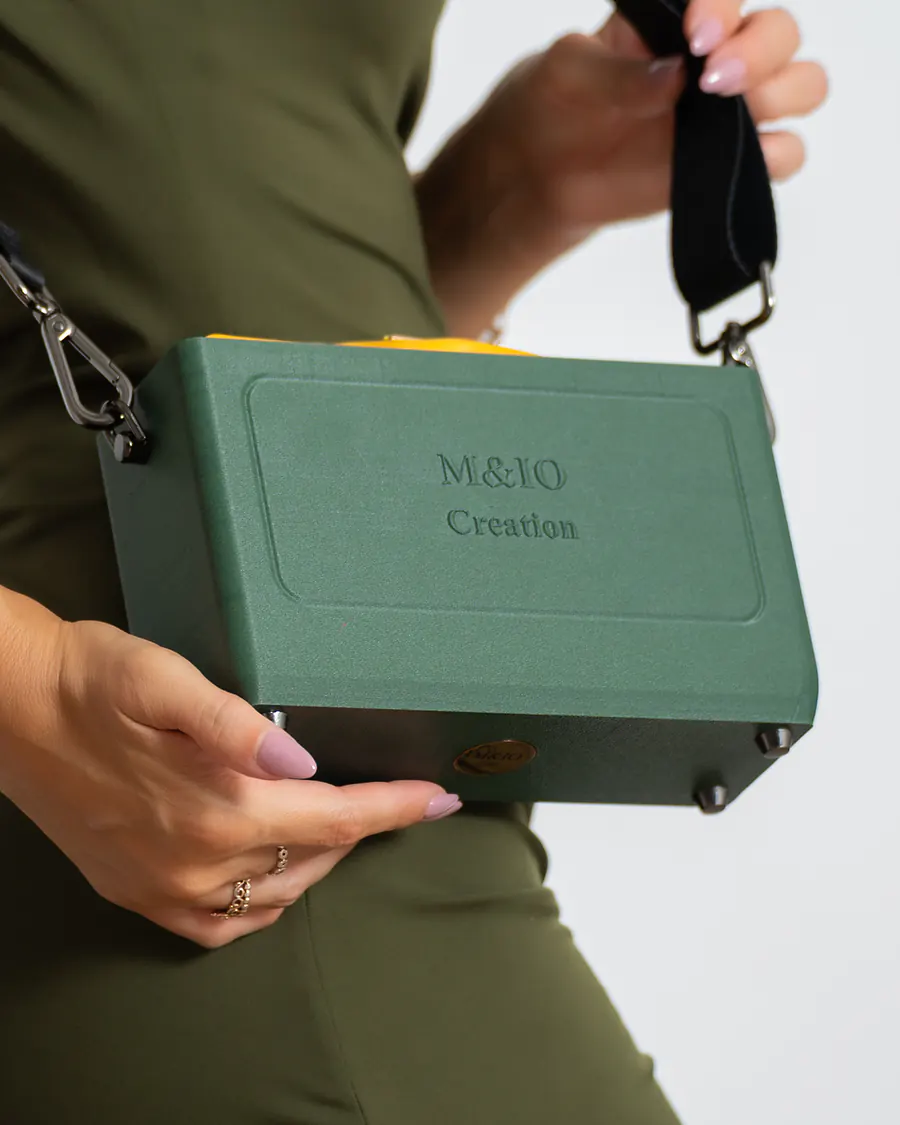 Da vicino, la borsa Ulivi di Puglia tenuta davanti al fianco, la cinghia nera tirata verso la spalla: sul fronte la scritta M&IO Creation in rilievo, dal bordo sporge la pochette gialla.